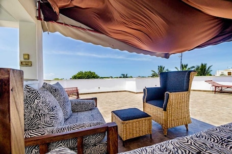 An outdoor seating area with wicker furniture and patterned cushions, a covered section, and a view of the beach.