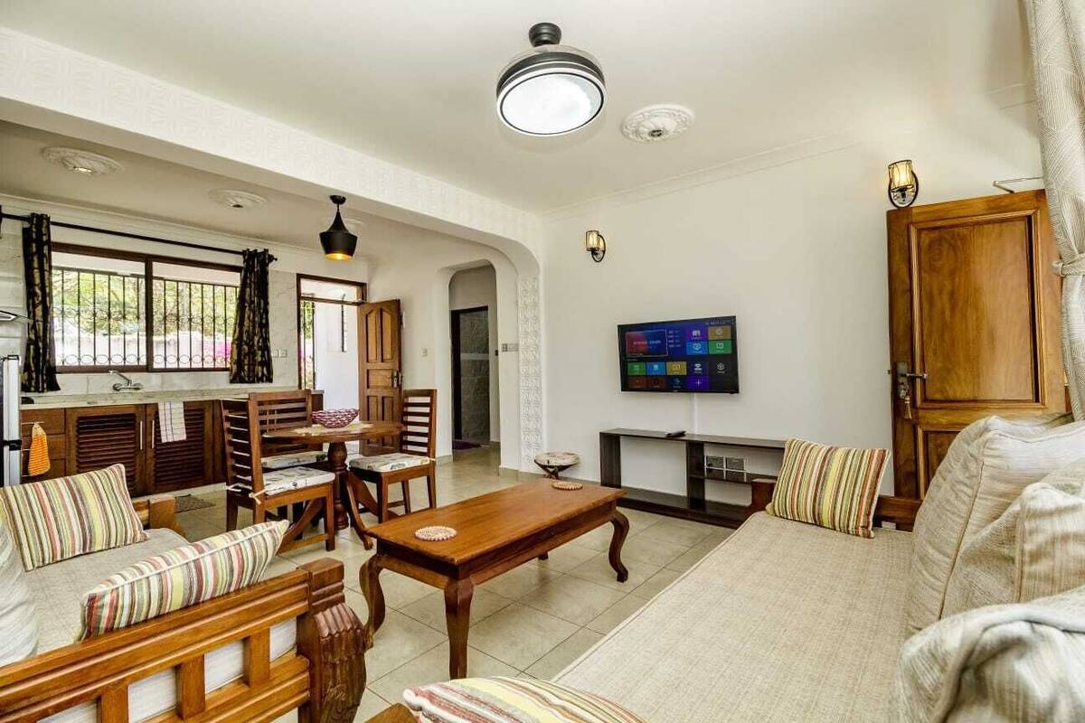 A spacious living area with a wooden dining set, a sofa, a TV mounted on the wall, and a wooden cabinet.