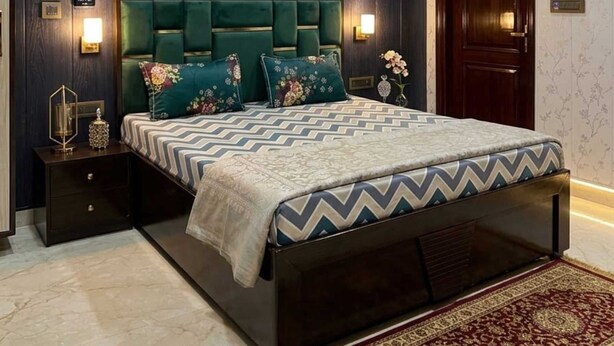 A bedroom with a dark wood panel wall, a green upholstered headboard, a patterned bedspread, a nightstand with a lamp, and a red patterned rug. - Hotel Shree Sanwaliya Inn (Ayodhya)
