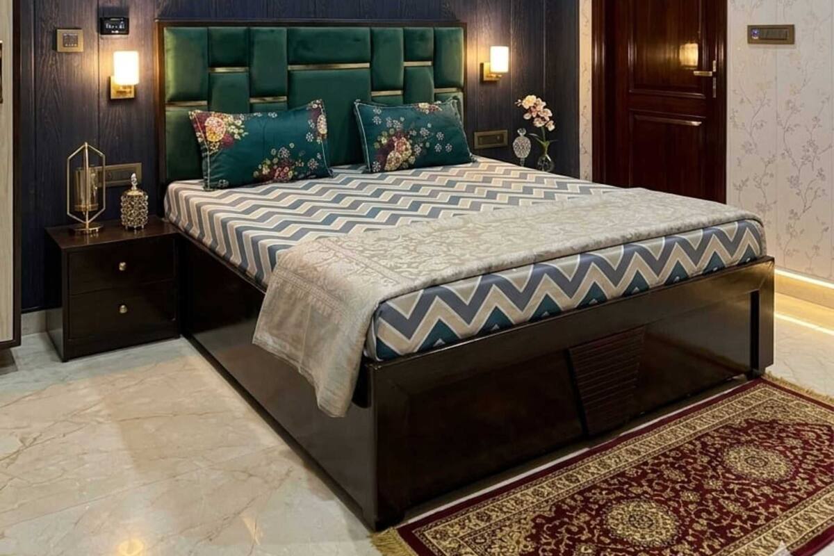 Un dormitorio con pared revestida de madera oscura, cabecera tapizada en verde, colcha estampada, mesita de noche con lámpara y alfombra roja con diseño.
