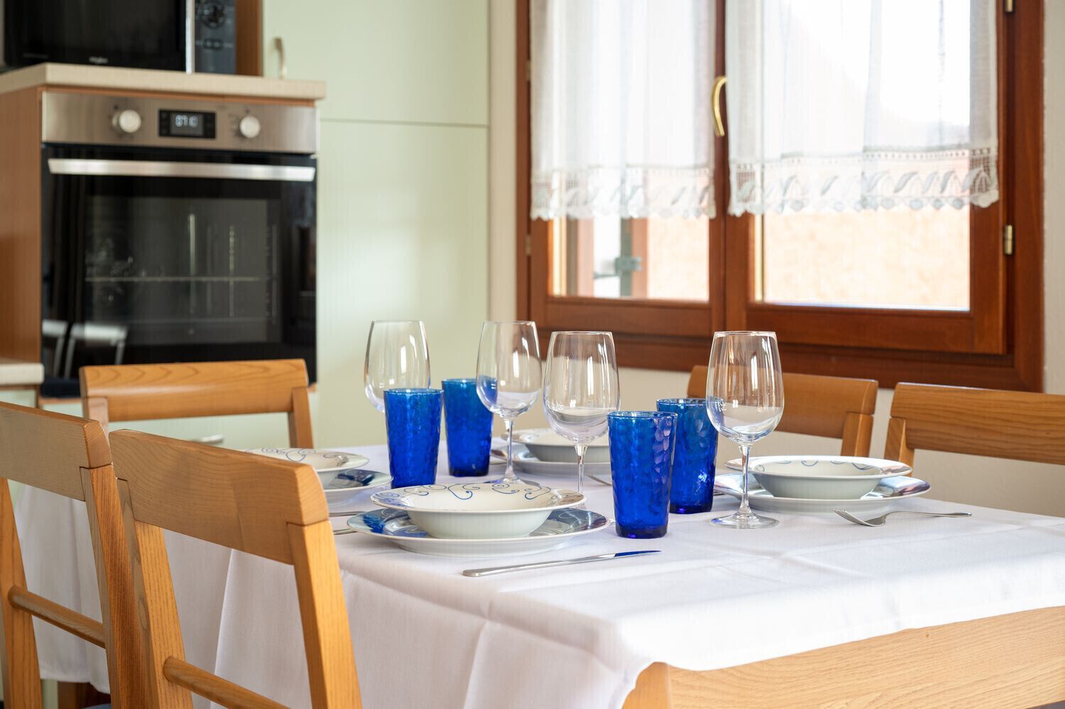 A dining table set with plates, glasses, and cutlery, with a kitchen in the background.