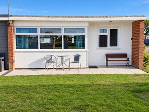 Ein eingeschossiges Gebäude mit einer Terrasse, die einen Tisch, zwei Stühle, eine Bank und ein Fenster mit Vorhängen zeigt.