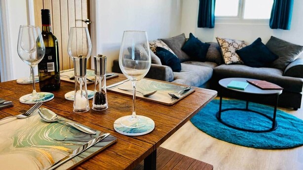 A dining table set with wine glasses, a bottle, and cutlery, a sofa, a coffee table, and a window with curtains. - Luxury Scratby chalet with free swimming (Great Yarmouth)