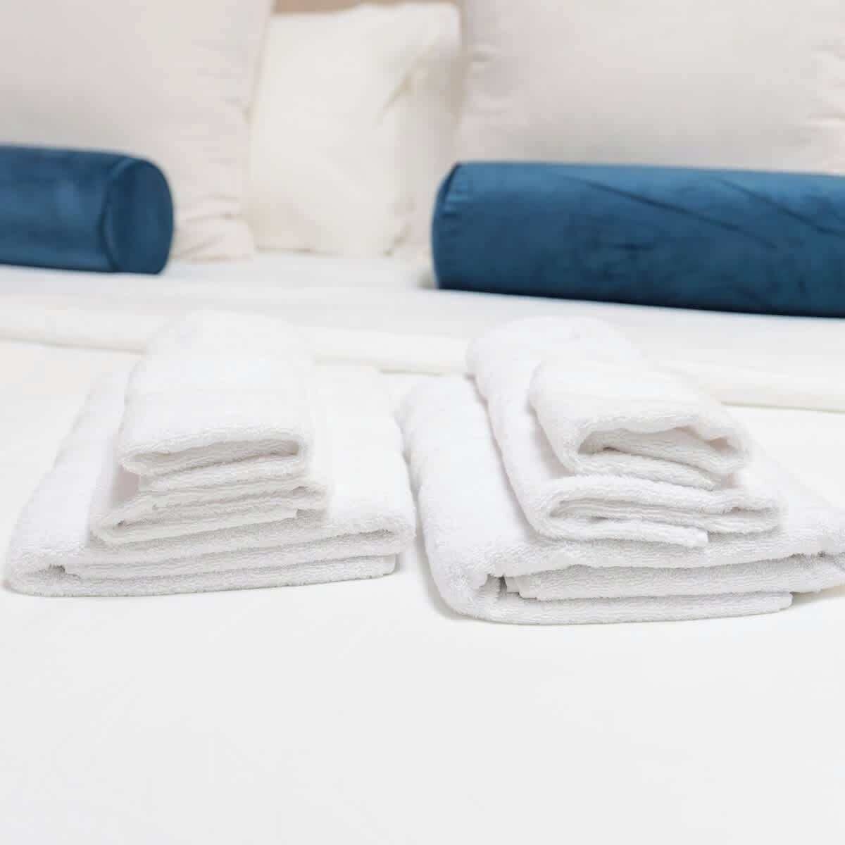 Una cama bien hecha con ropa de cama blanca y azul, y una pila de toallas blancas sobre la cama.