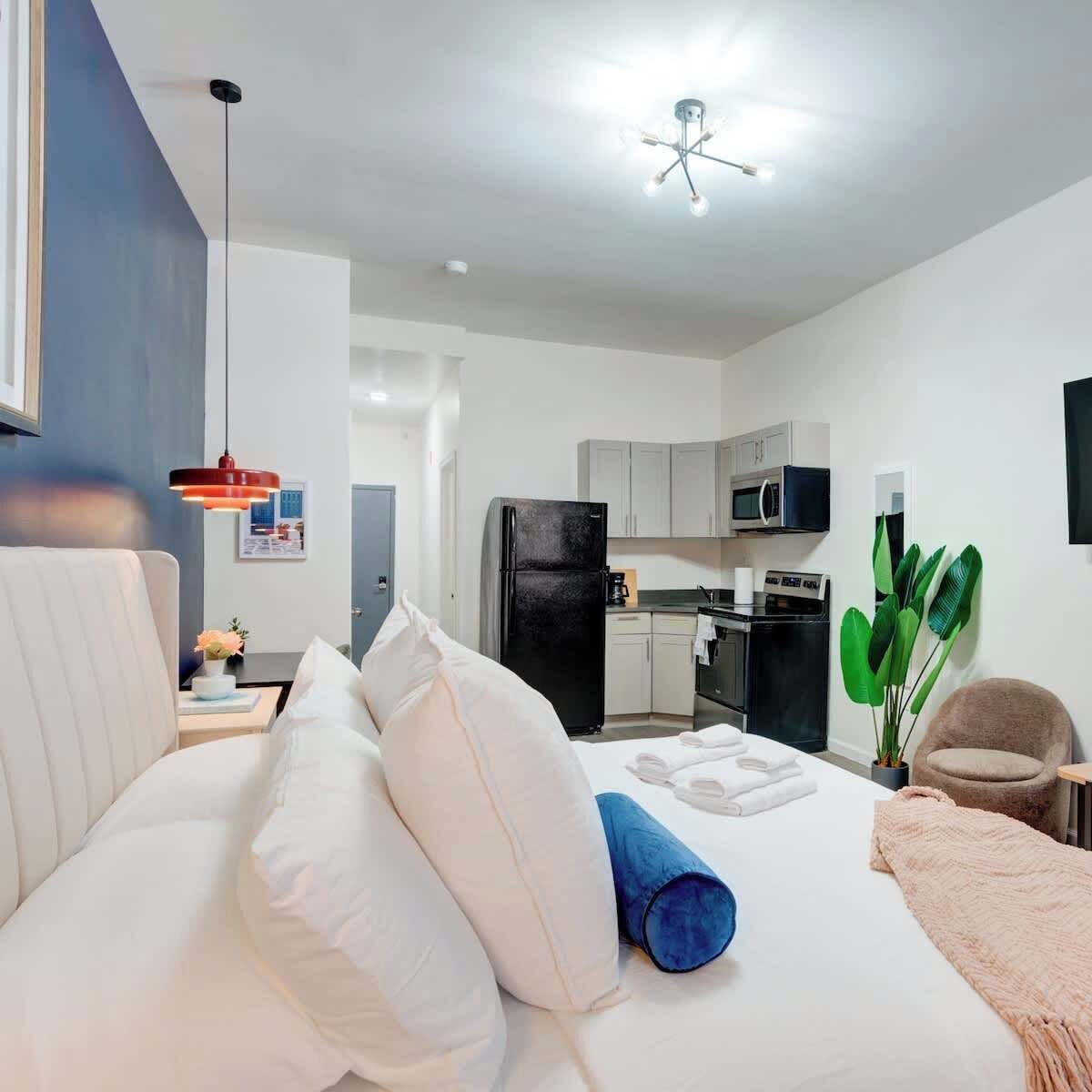 A modern hotel room with a bed, a desk, a chair, a kitchenette, and a ceiling light fixture.
