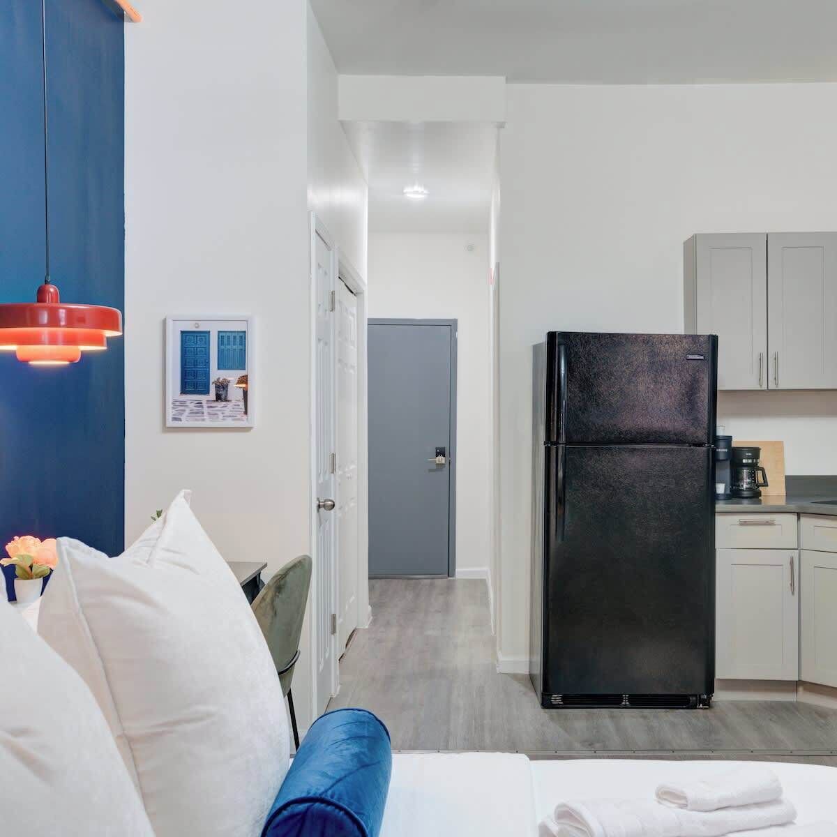 Une chambre d’hôtel compacte comprenant un lit, un mur d’accent bleu, une kitchenette avec un réfrigérateur et une porte donnant sur une autre pièce.