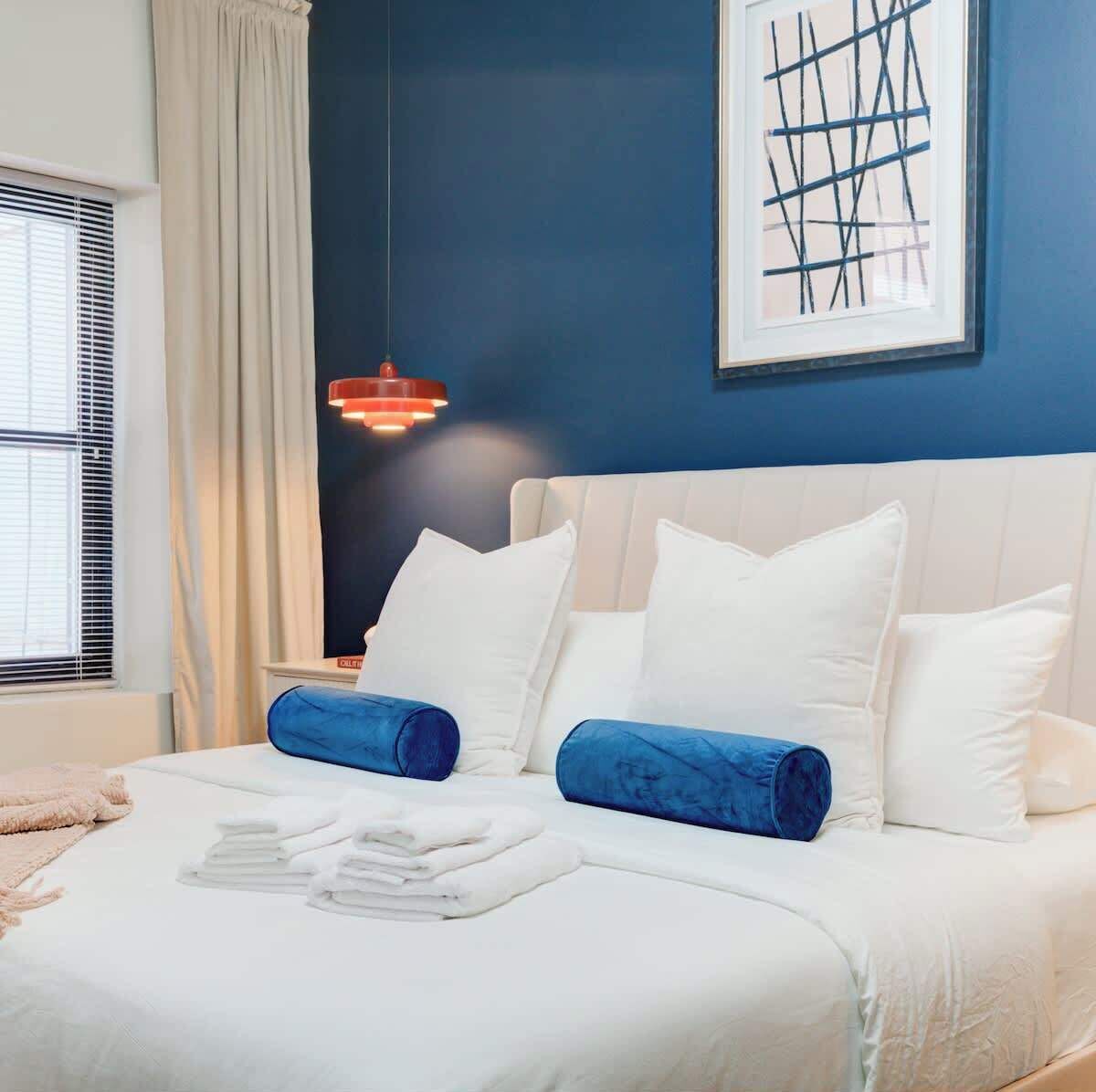 A neatly made bed with white and blue bedding, a blue wall, and a framed abstract artwork.