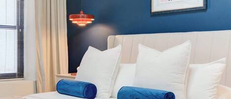 A neatly made bed with white and blue bedding, a blue wall, and a framed abstract artwork.