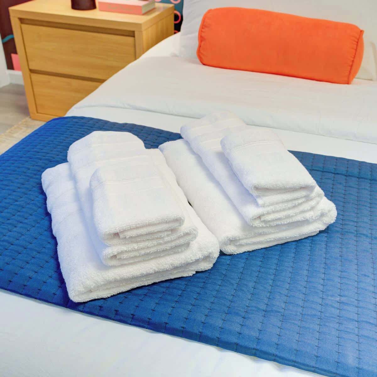 Ein ordentlich gemachtes Bett mit blauer, strukturierter Bettdecke, einem Stapel weißer Handtücher und einem orangefarbenen Kopfkissen.