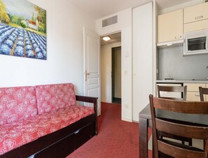 Ein Zimmer mit einem roten, gemusterten Sofa, einem Esstisch mit Stühlen, einer Küchenzeile mit einer Mikrowelle und einer Tür zu einem weiteren Raum.