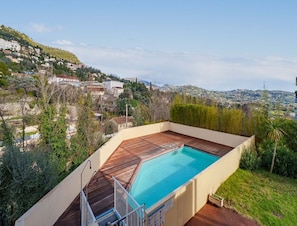 Ein Pool auf dem Dach mit einer Holzterrasse, umgeben von Grünpflanzen und mit Blick auf eine Stadt am Hang.