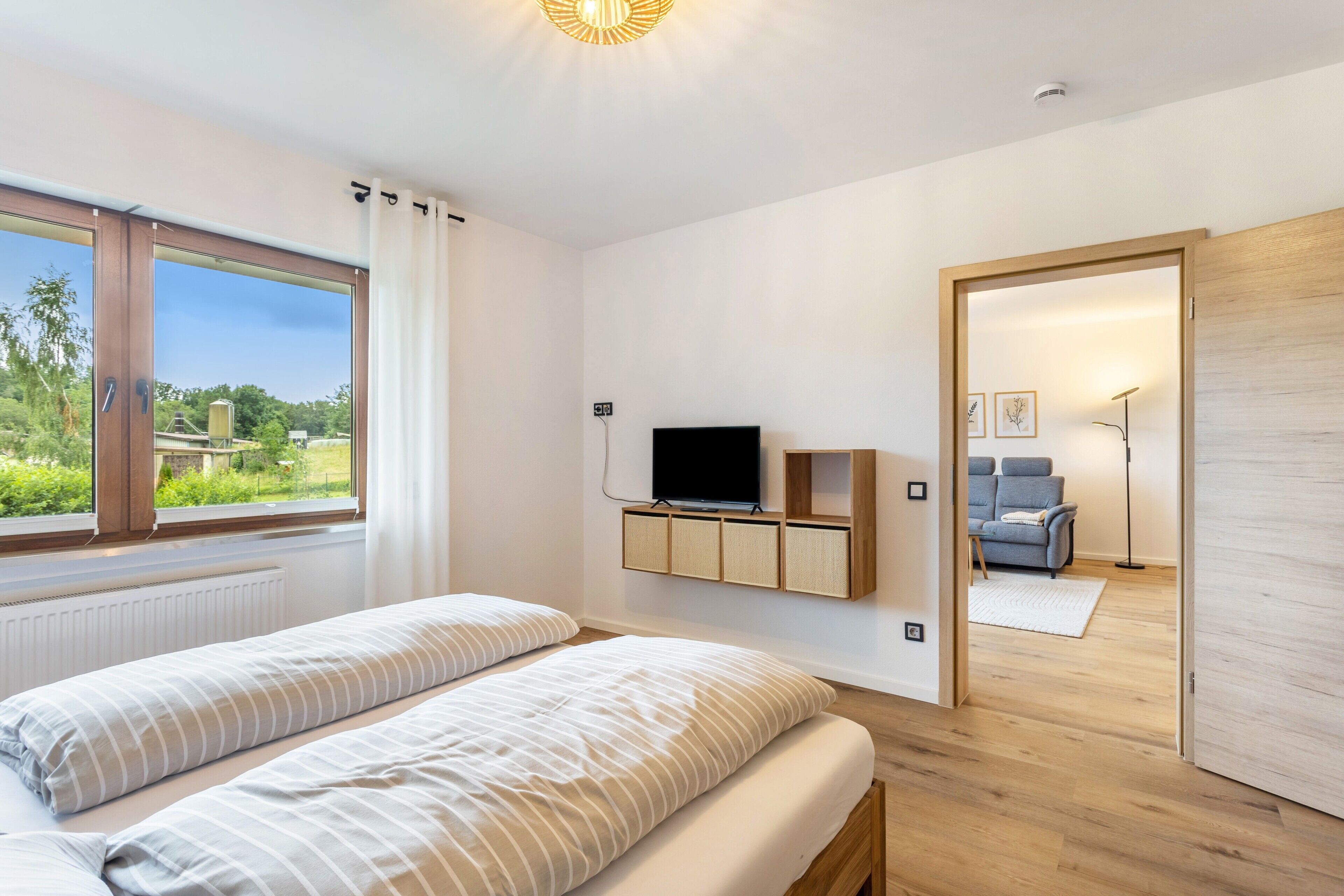 A bedroom with a bed, a TV, a wooden cabinet, and a view of greenery outside.