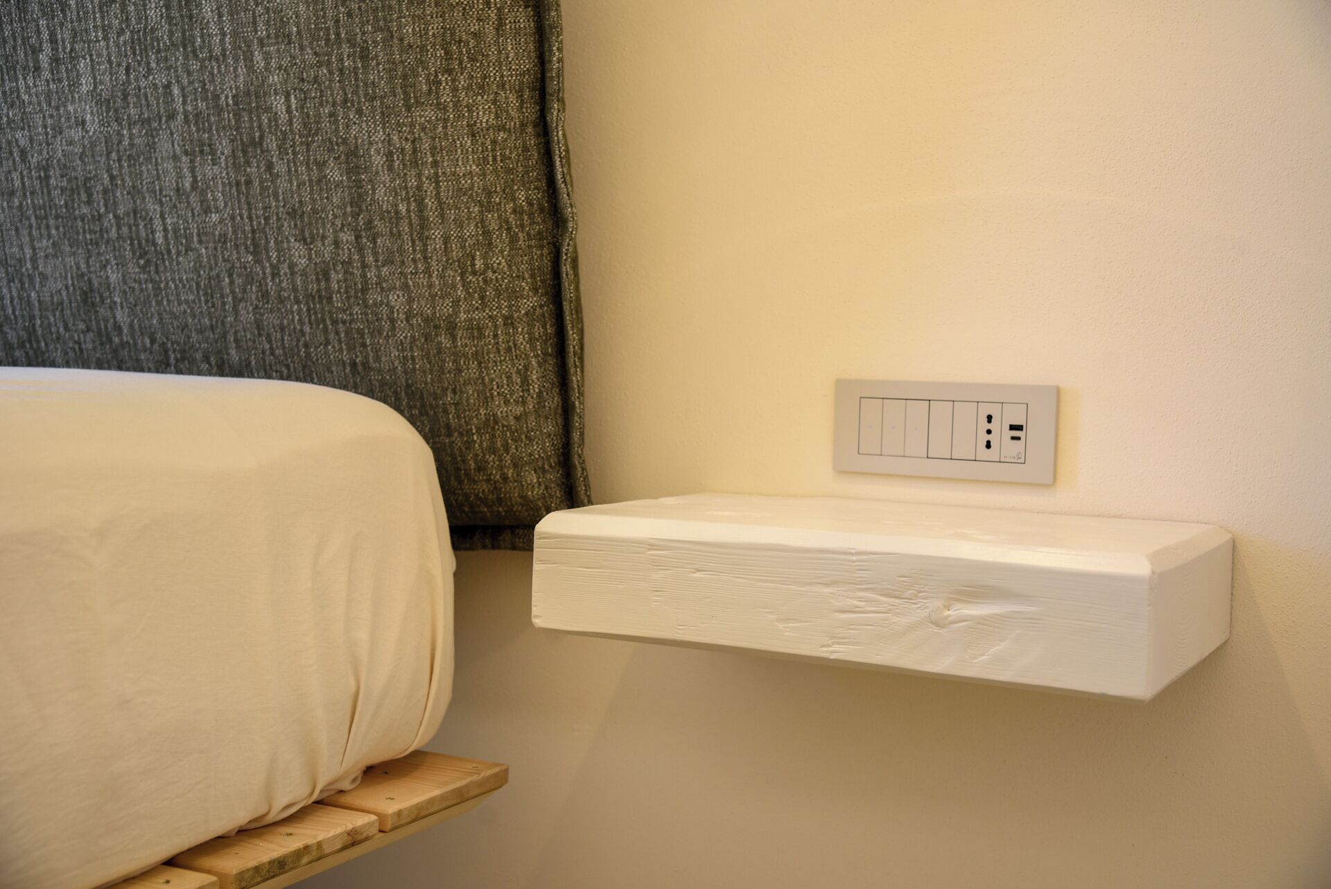 A bed with a white sheet, a wooden nightstand, and a wall-mounted electrical outlet.