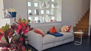 A modern living room with a sofa, colorful cushions, a potted plant, a unique ceiling design, and a staircase.