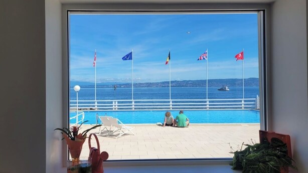 A view from a window overlooking a pool area with flags, a pier, and a distant cityscape.