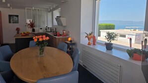 A modern kitchen with a dining area, a large window with a view of the sea, and a potted plant on the windowsill.