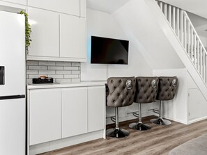 Eine moderne Küche mit weißen Schränken, einer schwarzen Arbeitsplatte und einem an der Wand montierten Fernseher. Drei braune, gepolsterte Barhocker mit Metallbeinen sind vorhanden.