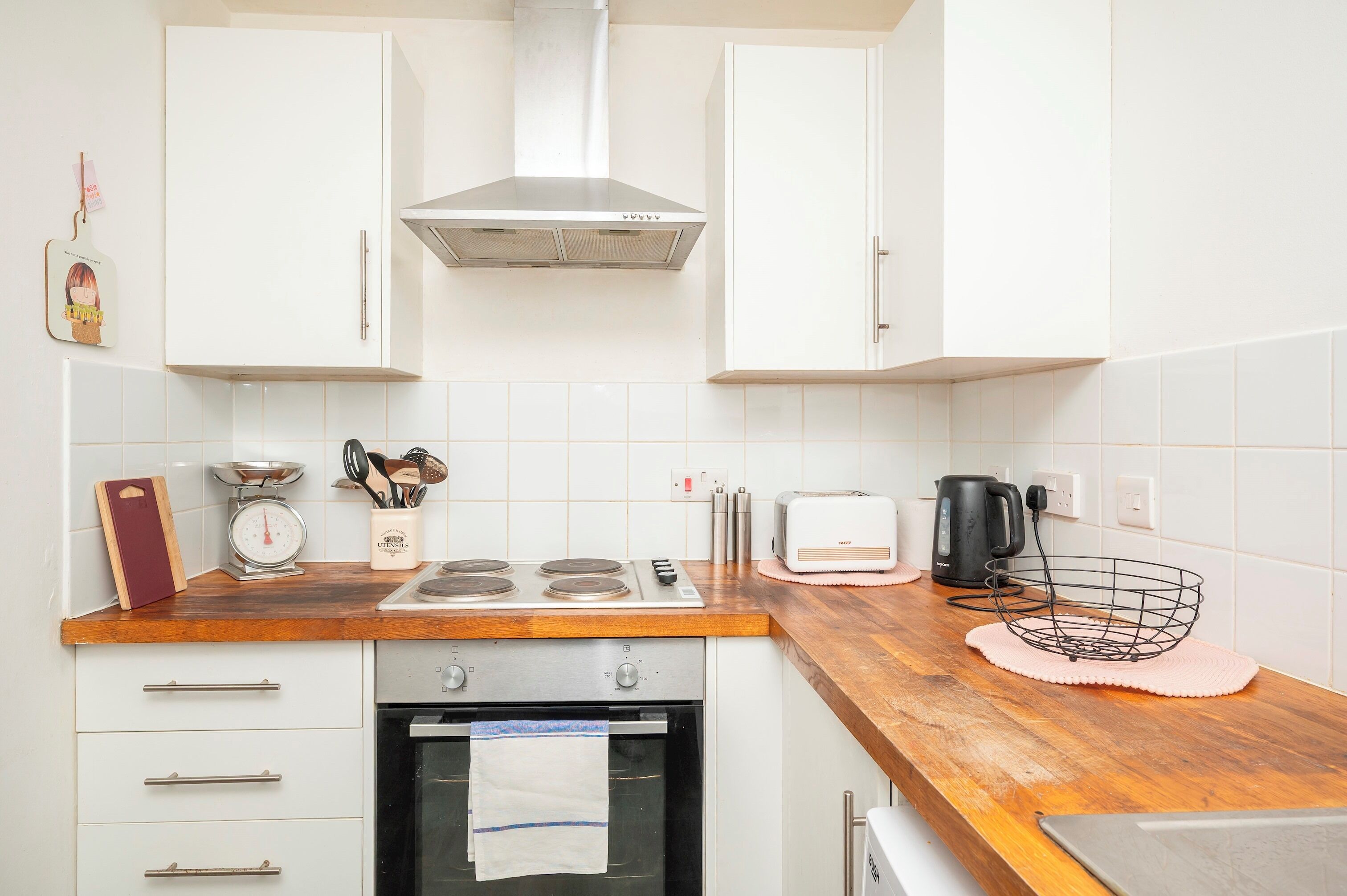 Eine Küche mit weißen Schränken, einer Edelstahl-Dunstabzugshaube, einer Holz Arbeitsplatte, einem Toaster, einem Wasserkocher und einem Drahtkorb.
