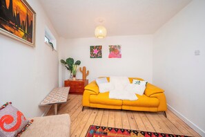 Ein Zimmer mit einem gelben Sofa, einem kleinen Tisch, einem gemusterten Teppich und zwei an der Wand befestigten Bildern.