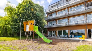 Ein Spielplatz mit Rutsche und Klettergerüst, ein Gebäude mit Balkonen und großen Fenstern, sowie ein Baum im Hintergrund.