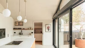 Een moderne keuken met een centraal eiland, hangende lampen en uitzicht door een glazen deur op een buitenruimte met een potplant.