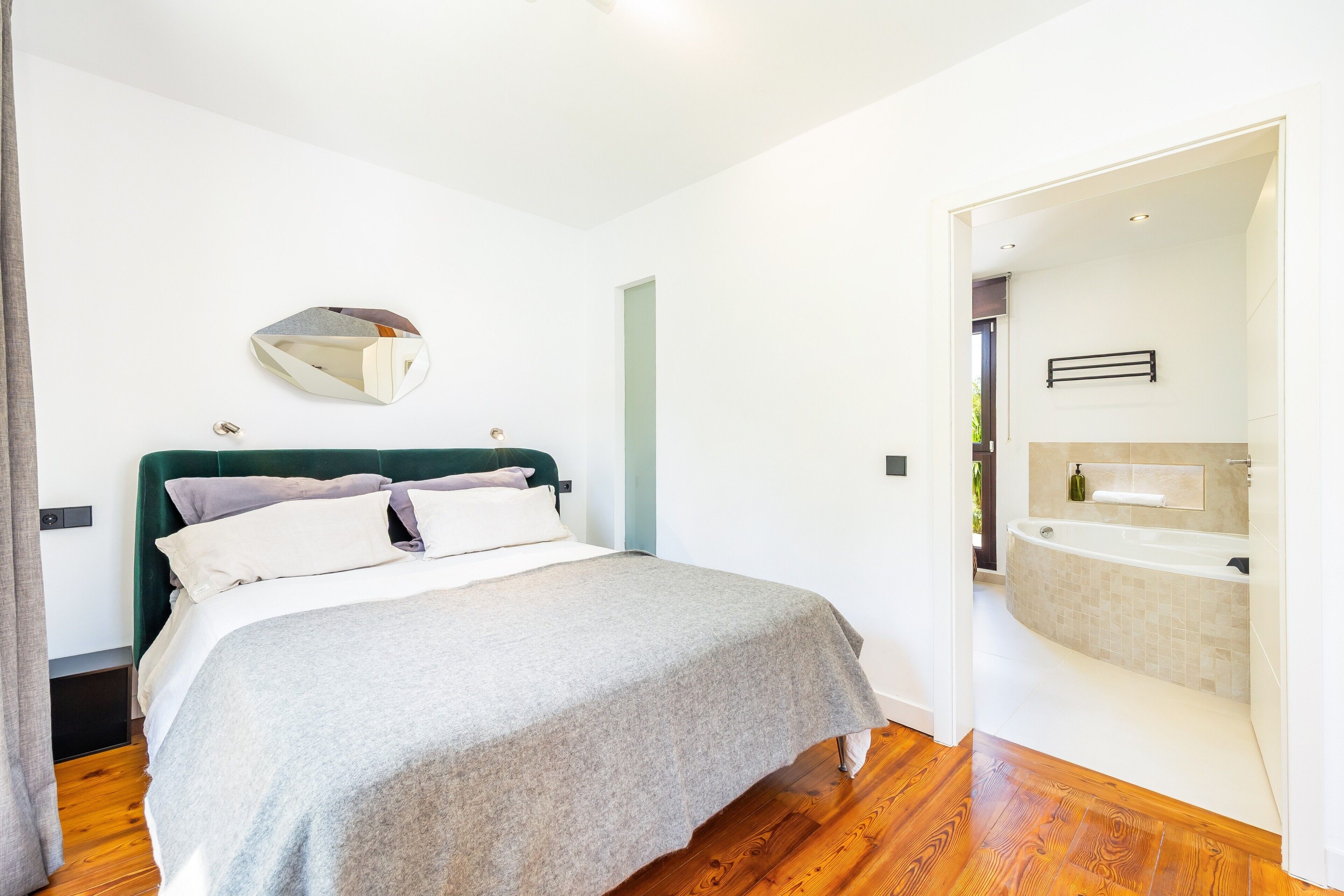 A bedroom with a bed, a bedside table, a mirror, and a bathroom with a bathtub.