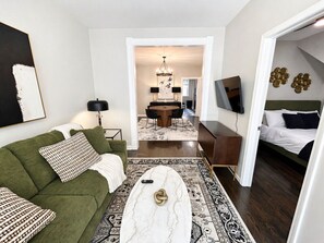 A living room with a green sofa, a coffee table, a TV, a dining area with a chandelier, and a bedroom with a bed. - Gothic-meets-mid-century 2BR in Lakeview | Statment Kitchen + Dining for 4 (Chicago)