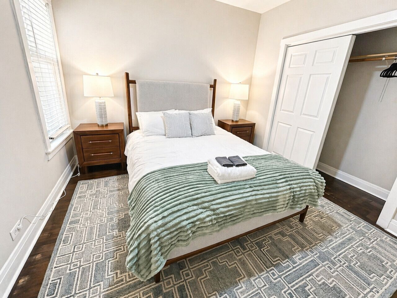 Ein ordentlich gemachtes Bett mit einer grünen Decke, zwei Nachttischlampen, einem Holz-Kopfteil und einem gemusterten Teppich.