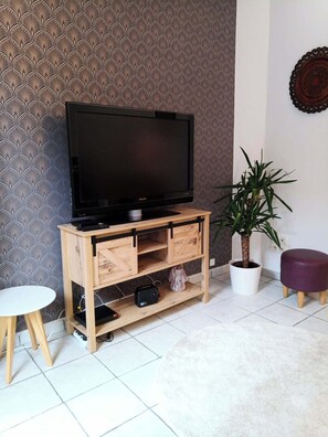 Un salon avec un téléviseur posé sur un meuble en bois, une plante en pot, une petite table ronde et un pouf violet.