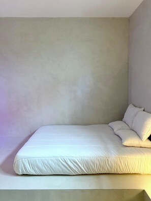 Ein Einzelbett mit weißer Bettwäsche in einem Raum mit schlichten Wänden und einer geneigten Decke.