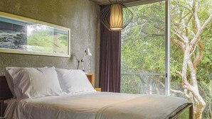 A bed with white linens, a framed picture on the wall, a bedside lamp, and a window with a view of trees.