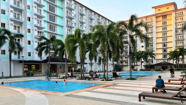 Zona de piscina con sillas de descanso, palmeras y un edificio de varios pisos al fondo.