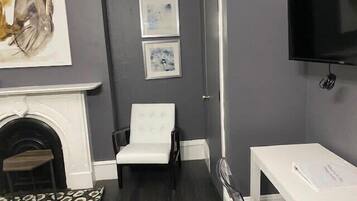 Ein Zimmer mit Kamin, weißem Ohrensessel, kleinem Tisch und Wand-Fernseher.