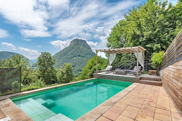 Ein Pool mit Bergblick, eine Pergola mit Stühlen und eine Holzwand.