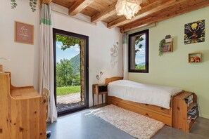 Ein Schlafzimmer mit einem hölzernen Bett, einem kleinen Nachttisch, einem Bücherregal und Blick ins Grüne durch eine Glastür.