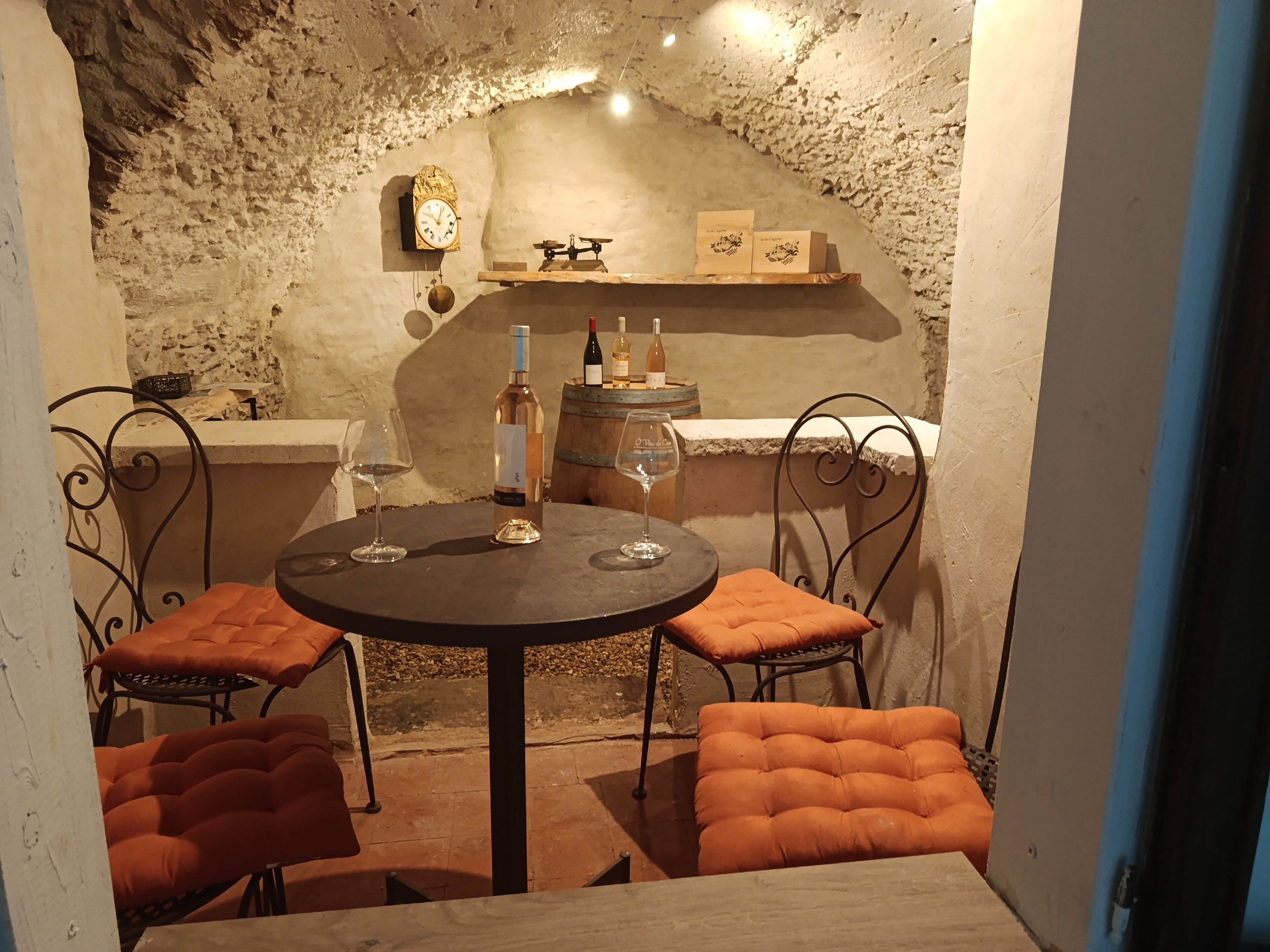 Una bodega rústica con una mesa redonda, dos sillas, botellas de vino y un reloj en la pared.
