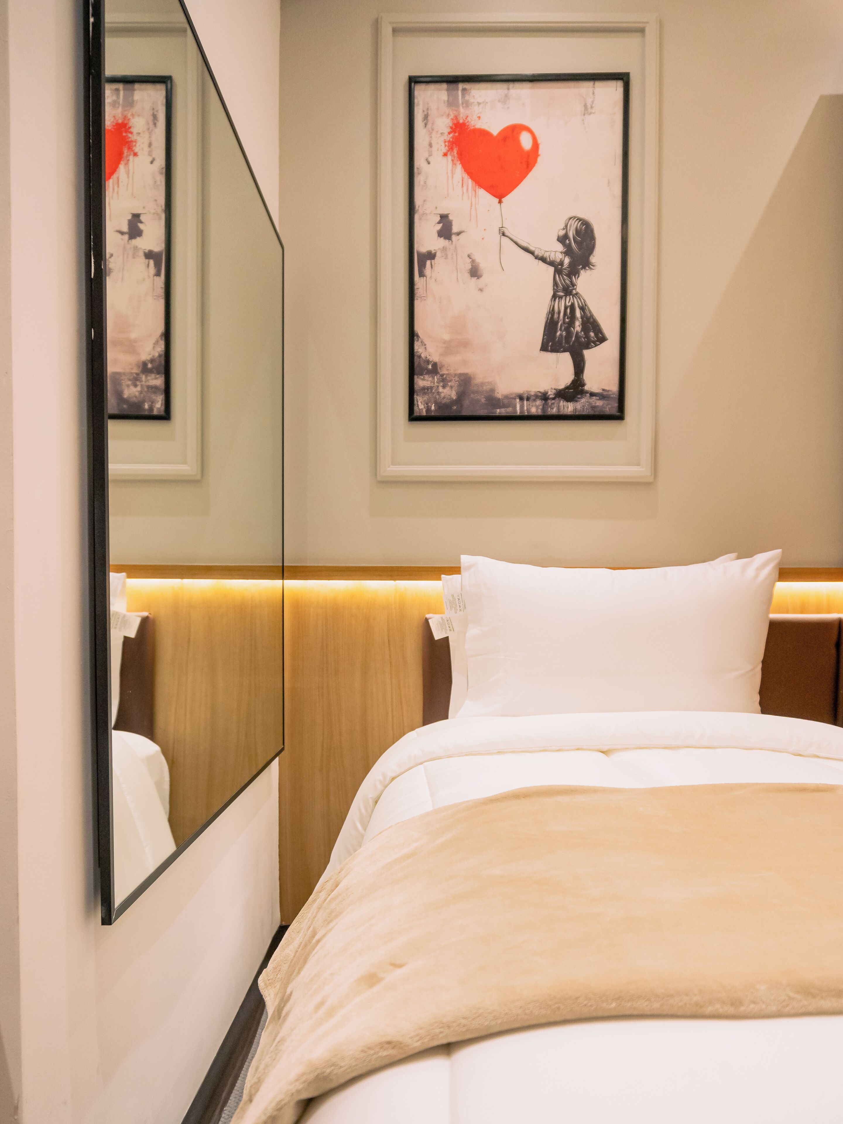 A hotel room with a bed, a large mirror, a framed artwork of a girl holding a heart, and a wooden headboard.