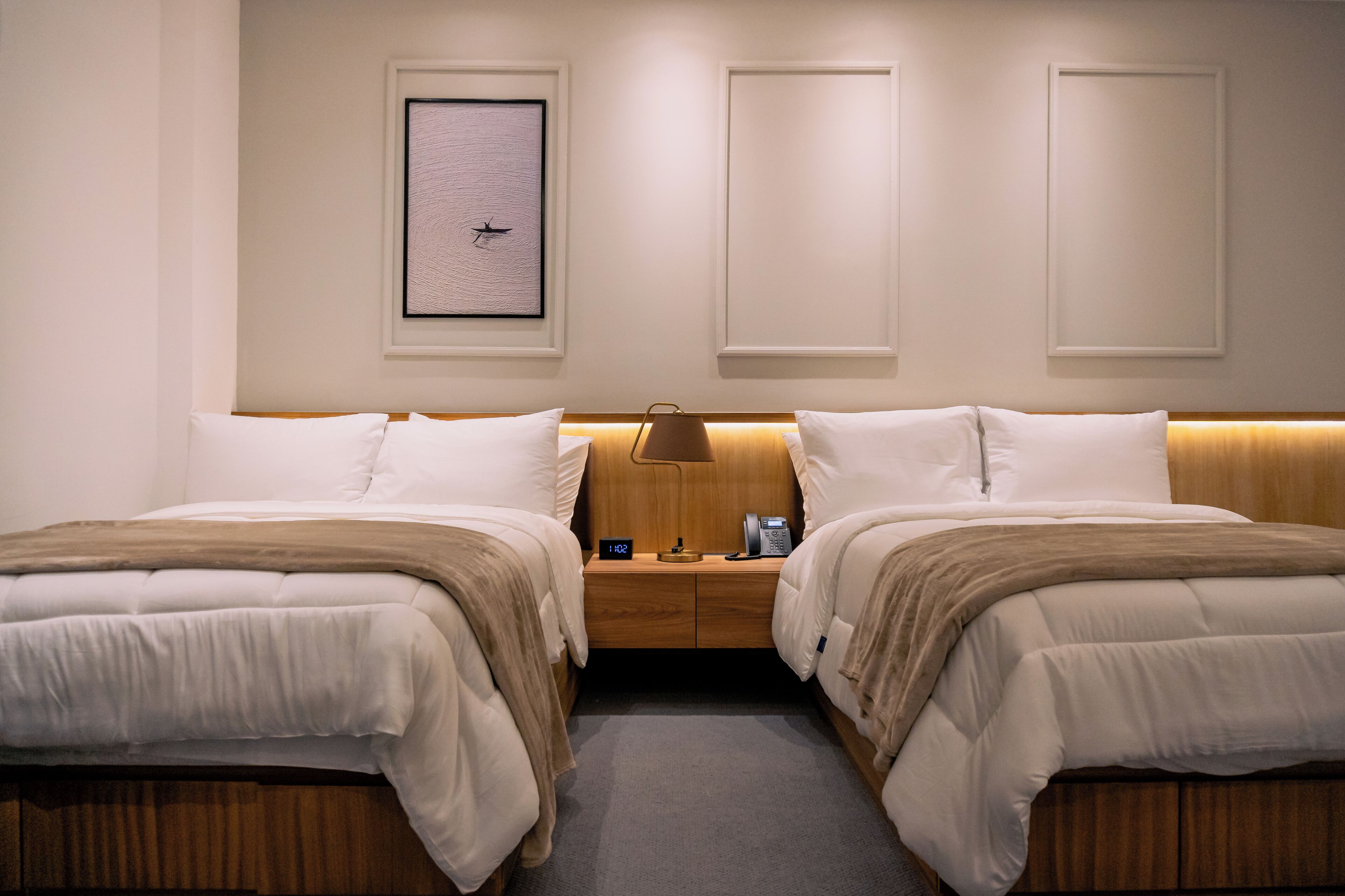 Hotellihuone, jossa on kaksi sänkyä, yöpöytä lampun kanssa, seinällä oleva kehystetty kuva ja kello.