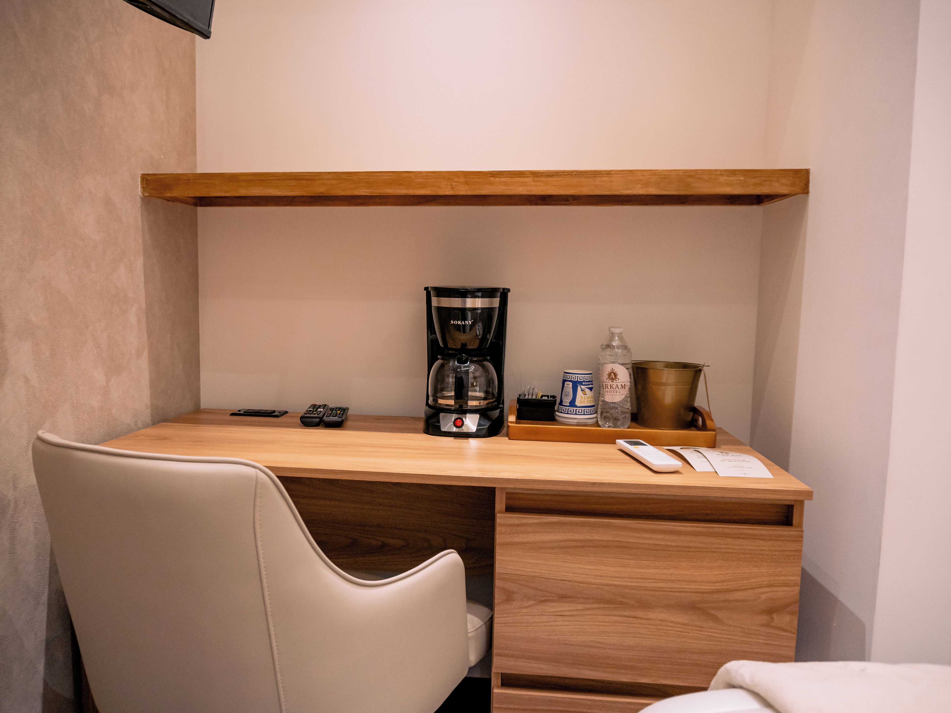 Una habitación de hotel pequeña con un escritorio de madera, una silla, una cafetera, una botella de agua, un cubo y un control remoto.