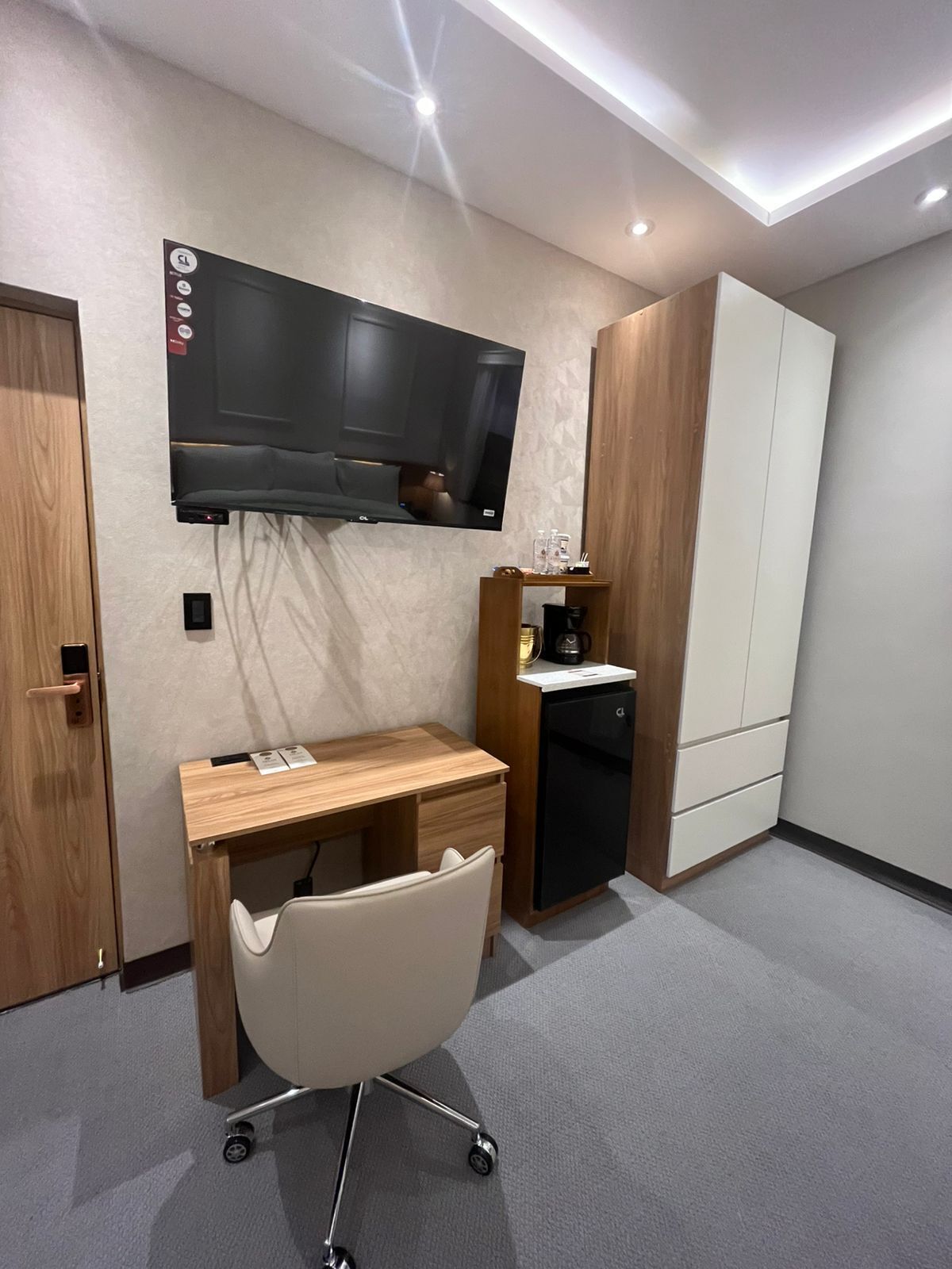 Habitación de hotel con televisor de pantalla plana montado en la pared, un escritorio de madera con silla, cafetera y armario.