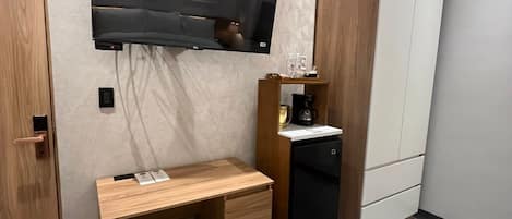 Habitación de hotel con televisor de pantalla plana montado en la pared, un escritorio de madera con silla, cafetera y armario.