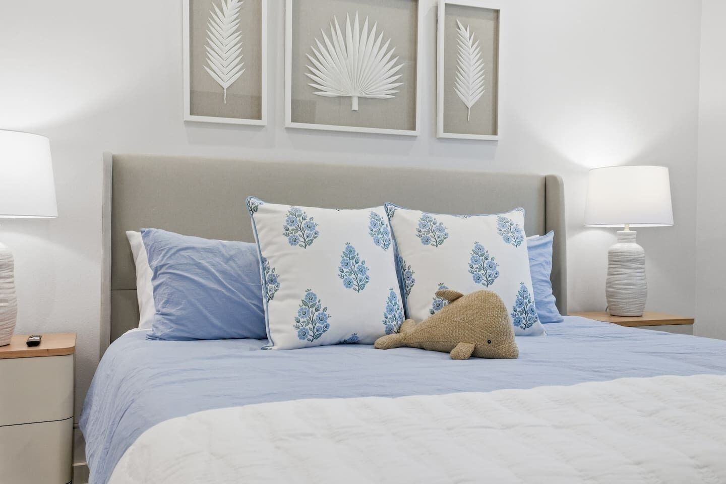 A neatly made bed with blue and white bedding, two pillows with a floral pattern, a brown stuffed animal, and three framed leaf designs on the wall.