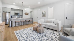 Una cocina moderna con una isla central, un comedor con sofá blanco y una sala de estar con sofá gris.