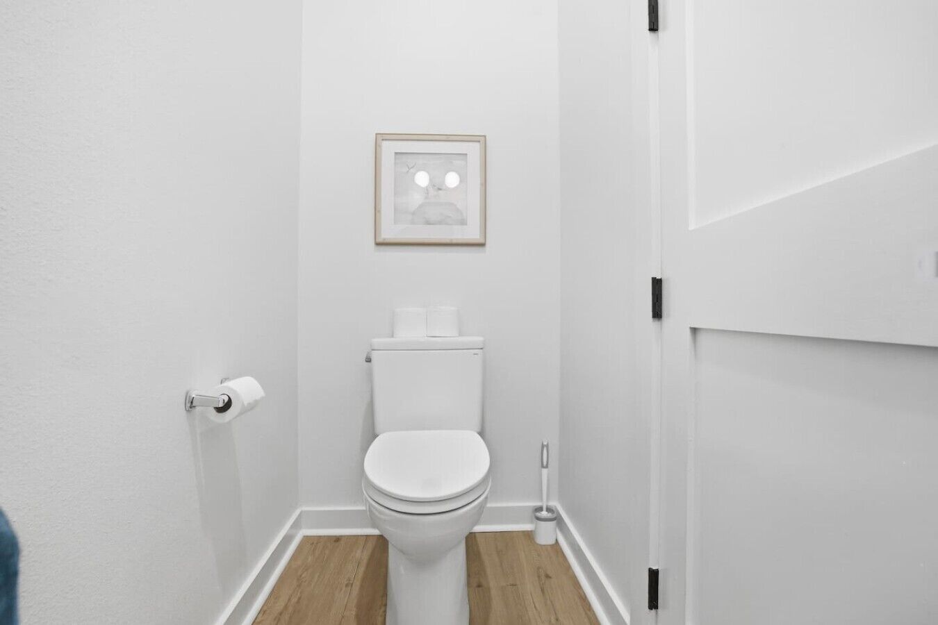 A small, white bathroom with a toilet, a framed picture on the wall, and a wooden floor.