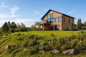 A modern two-story house with a large balcony, surrounded by a grassy landscape and trees. - Spa & Nature | L’Étreinte du Fleuve et des Cimes (Petite-Rivière-Saint-François)