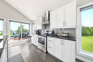 A modern kitchen with white cabinets, stainless steel appliances, and a large window overlooking a green landscape. - Spa & Nature | L’Étreinte du Fleuve et des Cimes (Petite-Rivière-Saint-François)