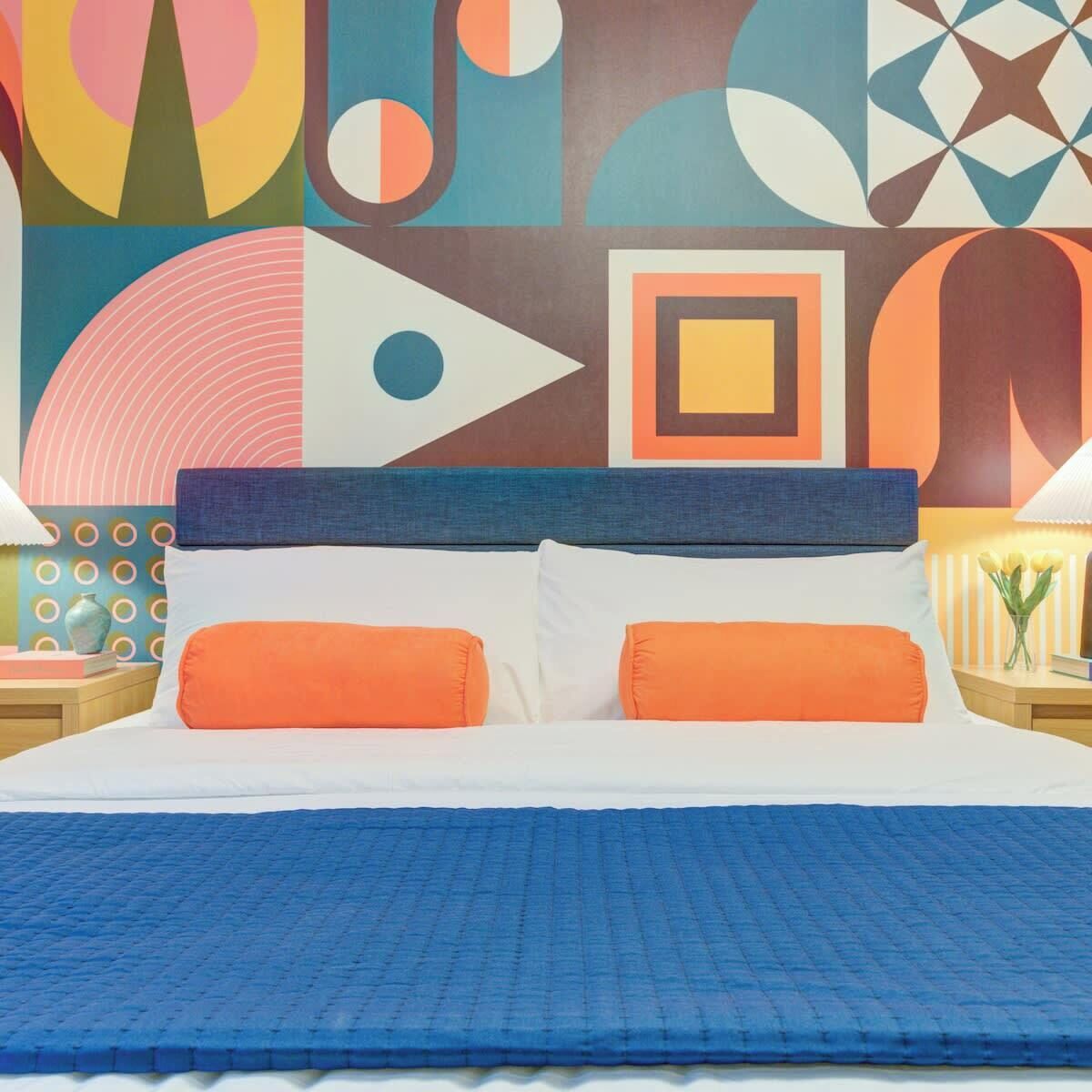 Una cama con cabecera azul, almohadas blancas y anaranjadas, y colcha azul.
