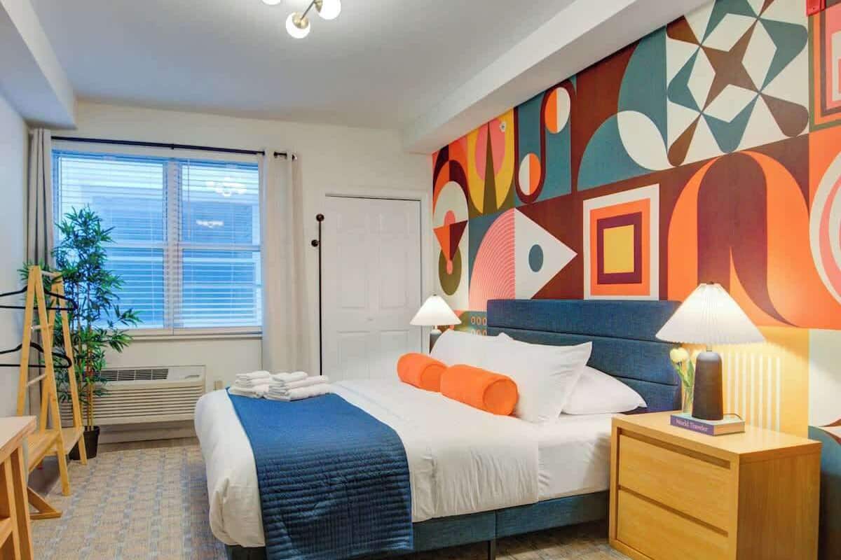 A modern hotel room with a large bed, a nightstand, a desk, and a colorful abstract wall mural.