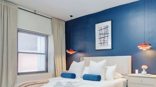 Une chambre à coucher moderne, avec un lit, des tables de chevet, une fenêtre avec des rideaux et une œuvre d’art accrochée au mur.
