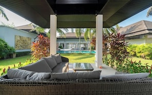 A covered outdoor seating area with a sofa and table, a pool in the background, and lush greenery. - Villa Lotus Heaven -Private 4BR Pool Villa. Phuket, Laguna area (Tambon Choeng Thale)